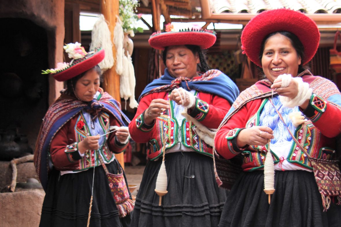 Three Indigenous women in Peru smile and hold spools of woven cloth.