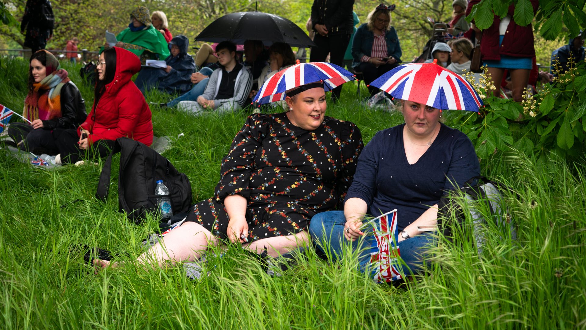 Two women wearing Union Jack umbrella hats sit in a park with others