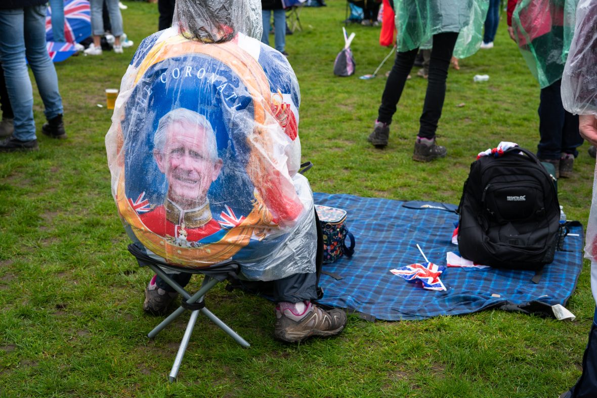 A man sat on a low picnic chair pictured from the back with a King Charles III coronation image on his back.