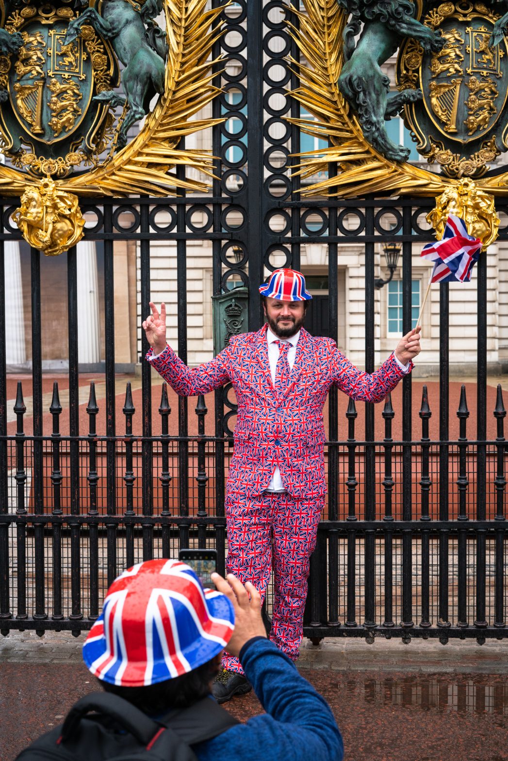 A man wearing a head-to-toe Union Jack suit, hat and umbrella photographed outside the gates of Buckingham Palace.