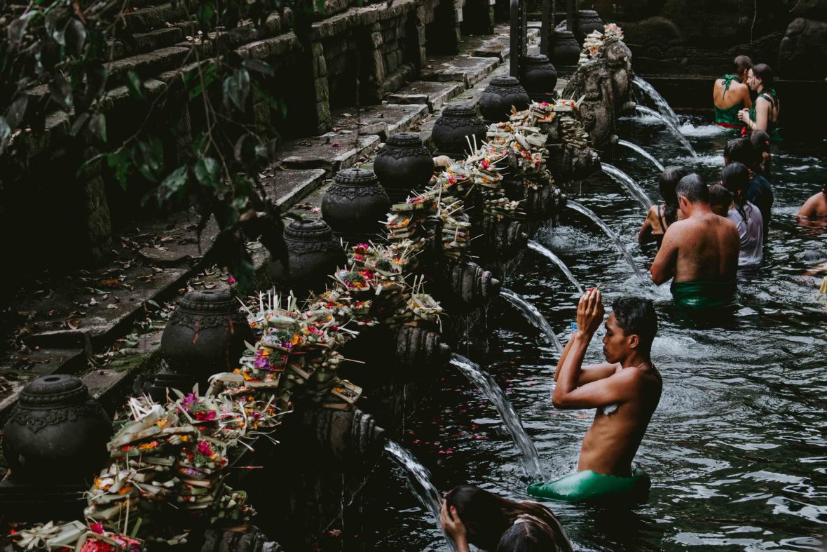 Men bathe in the waters at a holy temple. In the background are a couple of female tourists.