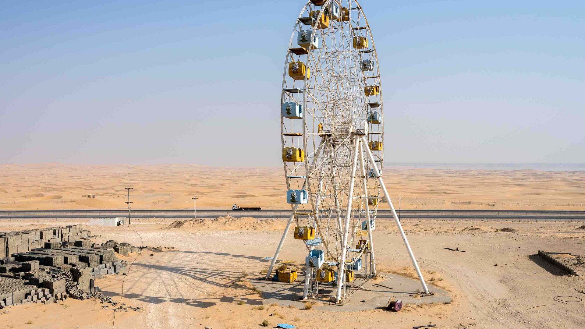 A ferris wheel stands in the desert with some buildings to its left.